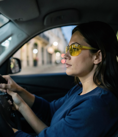 Lunettes pour la conduite de nuit et de jour – opticien à Boulazac Femme au volant portant des lunettes de conduite à verres jaunes dans une ville animée.